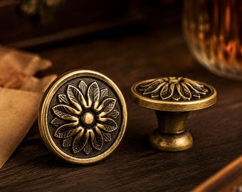 Può includere: Due pomelli rotondi per armadietti in metallo color ottone antico. Ogni pomello presenta un design floreale in rilievo e testurizzato con un elemento circolare centrale e petali radianti. Un pomello è visto frontalmente, l'altro di lato.