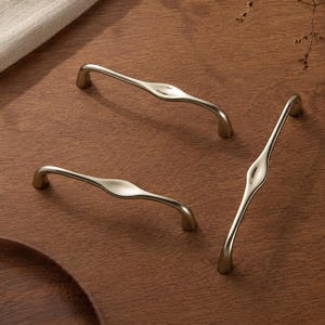 May include: Three metallic cabinet pulls with a smooth, curved, and elongated design. Each handle features a polished, reflective finish with a central bulge that tapers towards the ends, creating a fluid, organic shape for furniture hardware.