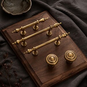 Può includere: Tre maniglie per armadietti in ottone antico con terminali decorativi e due pomelli rotondi abbinati. La ferramenta presenta una finitura metallica spazzolata ed è montata su una tavola di legno marrone scuro dal design classico.