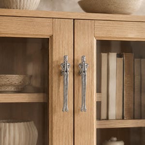 May include: Two silver-toned metal cabinet handles shaped like stylized human figures with elongated limbs. The figures are depicted in a seated position with hands touching their faces, mounted vertically on light oak wood cabinet doors.