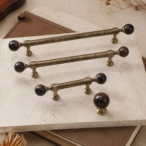 May include: A collection of three brass-toned metal drawer pulls of varying lengths and one matching round cabinet knob. Each piece features a brushed metallic finish and dark brown spherical ends, providing a consistent look for furniture hardware.