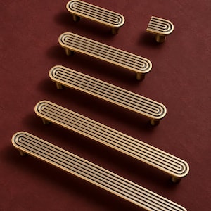 May include: A collection of six brass-toned metal cabinet pulls and one matching knob, each featuring a series of concentric, rounded rectangular grooves with dark recessed lines running along the length of the hardware pieces.