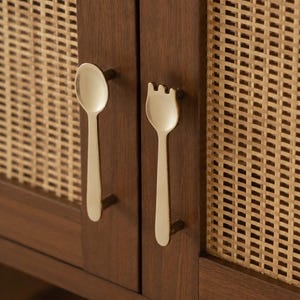 May include: A pair of gold-toned metal cabinet handles shaped like a spoon and a fork, mounted on dark brown wooden cabinet doors featuring light tan woven cane panels. The spoon handle is on the left and the fork handle is on the right.