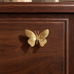 May include: A gold-colored metal cabinet knob shaped like a butterfly with detailed wing textures. The butterfly knob is mounted on a dark brown wooden drawer front, featuring a symmetrical design with two antennae and spread wings.