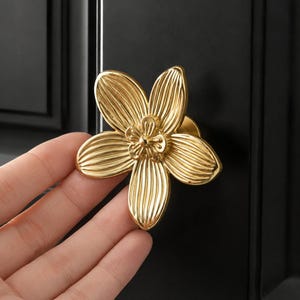 Puede incluir: Un pomo de mueble de metal dorado con forma de flor de cinco pétalos y un detalle floral central más pequeño. Los pétalos presentan texturas estriadas verticales y un acabado pulido y reflectante, montado sobre una puerta de armario negra.