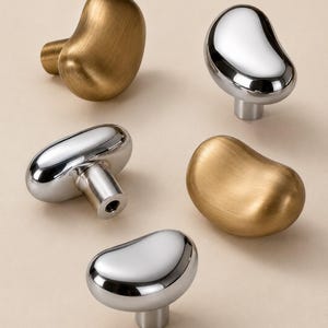 Op de afbeelding: Een verzameling van vijf kastknoppen met een onregelmatige, organische, boonachtige vorm. Drie knoppen hebben een gepolijste, reflecterende zilveren afwerking, terwijl twee knoppen een geborstelde, matte gouden afwerking hebben.