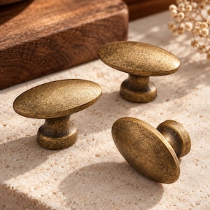 May include: Three oval-shaped cabinet knobs with a distressed antique brass finish. Each knob features a smooth, rounded top surface and a sturdy, tapered base, showcasing a weathered metallic texture with dark brown and gold tones.