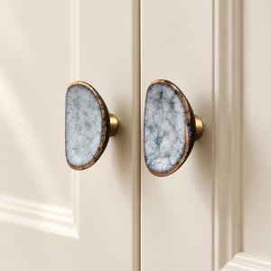 May include: Two semi-circular cabinet knobs featuring a textured, crackled white and grey ceramic face with a dark brown rim. Each knob is mounted on a cylindrical brass-toned metal base, attached to a pair of off-white cabinet doors.