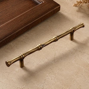 Op de afbeelding: Een messing kastgreep met een gestructureerd, op bamboe geïnspireerd ontwerp en een warme metallic afwerking. Het beslag heeft twee bevestigingspunten en een lange, slanke vorm die het gesegmenteerde uiterlijk van natuurlijke bamboestengels nabootst.