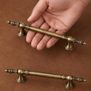 May include: Two antique brass cabinet pulls featuring a cylindrical bar design with decorative finials at each end. The hardware has a brushed metallic finish with dark highlights in the recessed areas, supported by two circular mounting bases.