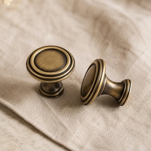 May include: Two round cabinet knobs with a brushed antique brass finish. Each knob features a circular face with a raised center and concentric ring detailing around the edge, supported by a tapered stem that flares at the base for mounting.