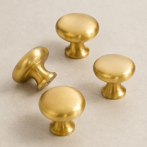 May include: Four round cabinet knobs with a brushed brass finish. Each knob features a smooth, domed top and a tapered base. One knob is positioned on its side, revealing the circular mounting hole at the bottom of the stem, while the others stand upright.