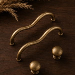 May include: Two antique brass cabinet pulls with a wavy, curved design and two matching round cabinet knobs. The hardware features a smooth, metallic finish with subtle darkened highlights, showcasing a consistent colour and texture throughout.