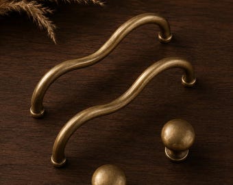 Può includere: Due maniglie per armadietto in ottone anticato con un design ondulato e curvo e due pomelli rotondi abbinati. La ferramenta presenta una finitura metallica liscia con sottili riflessi scuri, mostrando un colore e una consistenza uniformi.