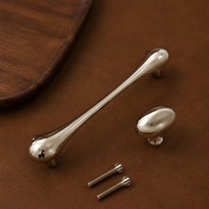 Peut inclure: Une poignée de meuble en métal argenté poli avec une forme incurvée et allongée, un bouton de meuble ovale assorti et deux vis de montage filetées en métal. La quincaillerie présente une finition lisse et réfléchissante.