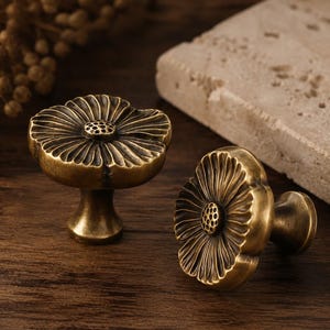 May include: Two brass cabinet knobs shaped like blooming flowers with detailed petals and a textured centre. The metal has a dark, antique finish with visible highlights on the edges of the petals and a sturdy, tapered base for mounting.
