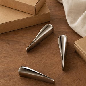 May include: Three polished silver-toned metal cabinet pulls with a tapered, elongated teardrop shape. Each handle features a smooth, reflective surface that curves gently to a point, providing a sleek and uniform appearance for furniture hardware.