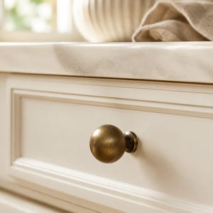 Peut inclure: Un bouton de meuble sphérique avec une finition en laiton antique brossé, monté sur une façade de tiroir de couleur crème. Le bouton présente une surface lisse et arrondie ainsi qu'une petite base circulaire fixée au panneau du tiroir.
