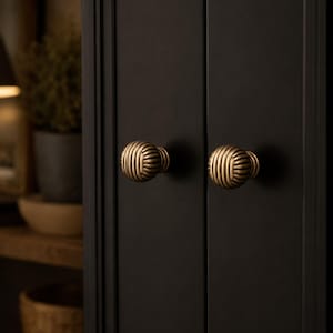 May include: Two round, brass-toned cabinet knobs with a ribbed, textured surface. Each knob is mounted on a matte black cabinet door with a vertical panel design. The knobs feature a series of parallel, raised lines that create a distinct pattern.