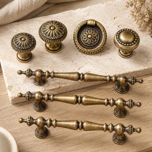 May include: A collection of antique brass-toned hardware including three long drawer pulls with decorative finials, two round floral knobs, one circular drop pull with a central flower motif, and one round knob with a beaded edge detail.