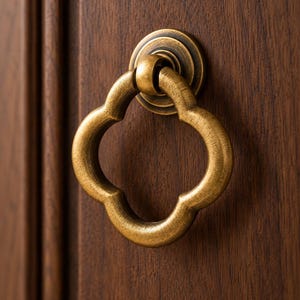 May include: A brass-toned metal ring pull with a quatrefoil shape, featuring a brushed finish and a circular mounting plate. The hardware is attached to a dark brown wooden surface with a visible grain pattern and a vertical panel edge.