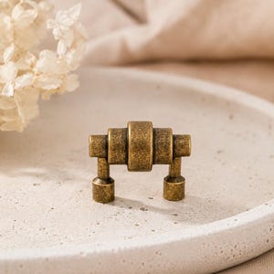 May include: A small, antique brass-toned metal cabinet pull featuring a horizontal cylindrical design with a central raised band and two vertical mounting posts. The hardware has a textured, weathered finish with visible dark patina accents.