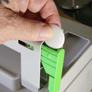 Op de afbeelding: Een witte ronde schijf wordt in een felgroen kunststof onderdeel geplaatst dat aan een groter wit frame is bevestigd. Het groene onderdeel heeft een verticale oriëntatie met zichtbare laagstructuren en een kleine cirkelvormige inkeping.