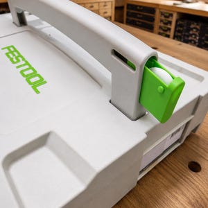 Può includere: Una custodia rettangolare grigio chiaro con una grande maniglia integrata e un meccanismo di chiusura verde brillante. La parola FESTOOL è stampata in lettere verde brillante sulla superficie superiore della custodia, che presenta una finitura in plastica testurizzata.