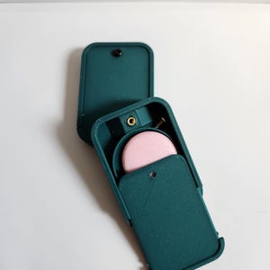 Puede incluir: Una caja rectangular de color verde azulado impresa en 3D con tapa abatible. El interior presenta un disco circular de color rosa claro y un pequeño inserto roscado de color dorado metálico. La caja tiene una superficie texturizada y un cierre negro.