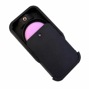 Könnte beinhalten: Eine schwarze, rechteckige Smartphone-Hülle mit einem verschiebbaren Kamera-Abdeckungsmechanismus. Die Hülle verfügt über einen kreisförmigen Ausschnitt für das Kameraobjektiv, der einen rosa Filter freigibt, sowie einen messingfarbenen Gewindeanschluss.