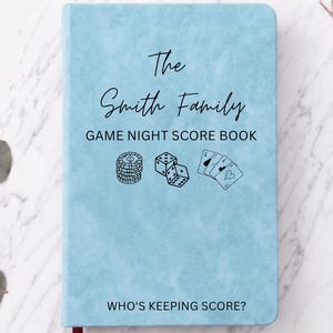 Peut inclure: Un carnet de score bleu clair avec un marque-page ruban rouge foncé. La couverture porte le texte "The Smith Family GAME NIGHT SCORE BOOK" et "WHO'S KEEPING SCORE?" ainsi que des illustrations au trait noir de jetons, dés et cartes à jouer.