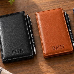 May include: Three rectangular pocket notebooks in black, tan, and navy blue, each featuring a textured cover with embossed initials and a matching black pen held in a side loop. The notebooks are arranged side-by-side on a wooden surface.