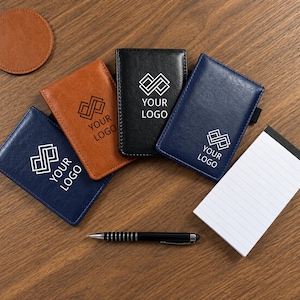 May include: Three rectangular card holders in navy blue, tan, and black, each featuring a white geometric logo and the text YOUR LOGO. A white lined notepad, a black ballpoint pen, and a circular tan coaster are arranged alongside the holders.
