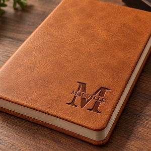 May include: A rectangular notebook with a textured tan brown cover. The front cover features a large embossed letter M with the word MANVILLE printed horizontally across the centre of the letter. The notebook has white pages visible along the edge.
