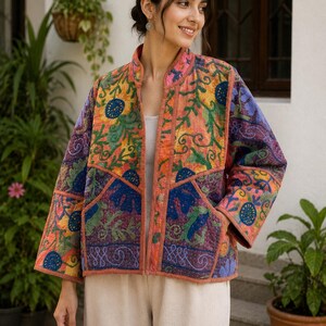 May include: A quilted jacket featuring a vibrant patchwork of floral and geometric embroidery in shades of orange, purple, blue, and green. The jacket has a mandarin collar, long sleeves, two front pockets, and a solid orange trim along the edges.