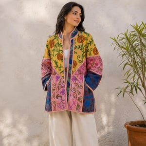 May include: A long-sleeved, open-front quilted jacket featuring a vibrant patchwork design with floral and pomegranate motifs in shades of yellow, pink, and blue. The garment has a mandarin collar and light pink trim along the edges and seams.