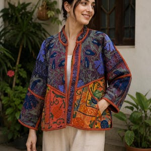 Könnte beinhalten: Eine gesteppte Jacke mit Patchwork-Design und lebendigen floralen sowie geometrischen Stickereien in Orange, Blau, Violett und Dunkelgrau. Die Jacke verfügt über einen Stehkragen, lange Ärmel und zwei Vordertaschen mit orangefarbenen Kanten.