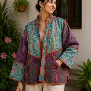 May include: A quilted jacket featuring a patchwork design in shades of teal, purple, and orange. The garment has a mandarin collar, long sleeves, two front pockets, and a rust-coloured trim along the edges, showcasing intricate floral embroidery.