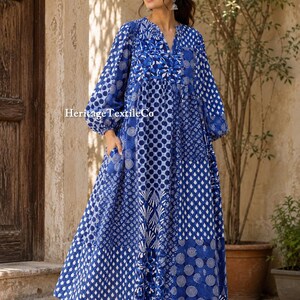 Puede incluir: Un vestido de manga larga hasta el suelo con un patrón de patchwork azul y blanco que presenta varios motivos geométricos y circulares. La prenda tiene cuello en V, mangas fruncidas y bolsillos laterales. El texto Heritage TextileCo es visible.