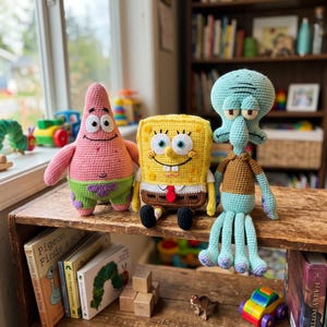 May include: Three crocheted plush toys representing cartoon characters: a pink starfish in green shorts, a yellow rectangular sponge with a white shirt and red tie, and a light blue cephalopod with a brown shirt, all featuring stitched details.