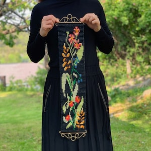 May include: A black woven wall hanging with a floral design in orange, yellow, green, and blue. The tapestry is rectangular with a gold-colored metal top and bottom. The floral pattern includes various flowers and leaves.