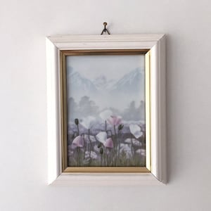 May include: A framed floral print featuring pink and white flowers in a field with a mountain backdrop. The artwork is set in a white frame with a gold inner border, hanging on a white wall.