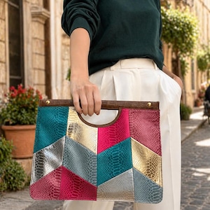 Peut inclure: Une pochette patchwork colorée avec une poignée marron. La pochette présente un motif géométrique en similicuir turquoise, argent, doré et rose. Le sac est tenu par une personne portant un pull vert foncé et un pantalon blanc.