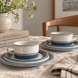 May include: Two ceramic tea cups and matching saucers featuring a cream-colored base with horizontal bands in shades of blue and tan. Each cup has a side handle and sits on a saucer with concentric circular stripes in the same color palette.