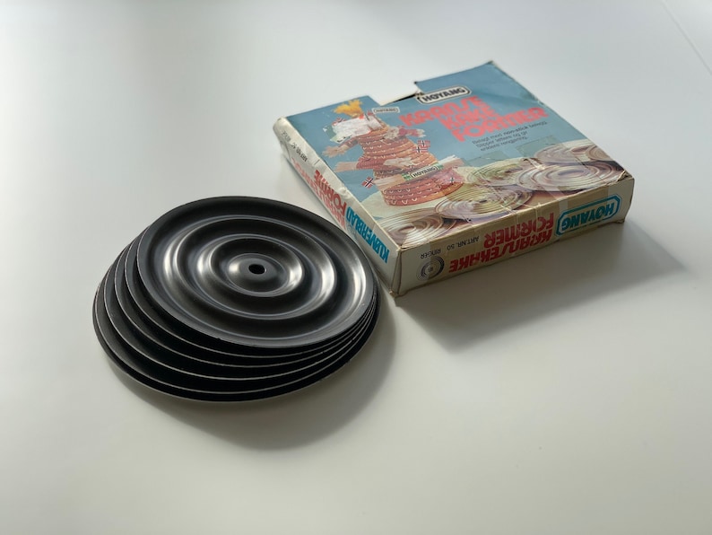 Op de afbeelding: Een set van zes zwarte metalen taartringen, gestapeld op elkaar. De ringen zijn ontworpen om gelijkmatige lagen voor taarten te cre&euml;ren. De doos voor de taartringen staat op de achtergrond en heeft de merknaam "ROYAL CAKE" en de tekst "Art. Nr. 30100".