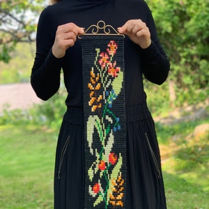 May include: A black tapestry with a floral design in shades of red, orange, green, and blue. The tapestry is rectangular with a gold-colored metal top and bottom. The floral pattern includes various flowers and leaves.