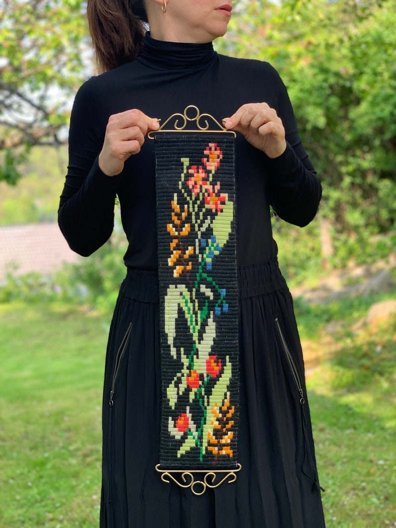 May include: A black tapestry with a floral design in shades of red, orange, green, and blue. The tapestry is rectangular with a gold-colored metal top and bottom. The floral pattern includes various flowers and leaves.