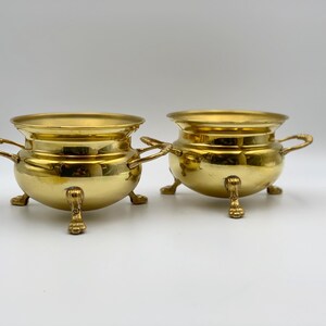 May include: Two antique brass pots with handles and claw feet. The pots have a rounded body and a wide rim. The brass has a polished, reflective surface, and the pots are set against a white background.