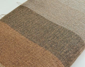 Puede incluir: Un textil doblado con una textura tejida, con rayas horizontales en tonos beige, marrón y bronceado. La tela parece ser una fibra natural, posiblemente lino o un material similar. Las rayas crean un patrón visual.