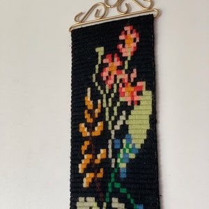 May include: A black tapestry with a floral design in shades of orange, pink, green, and blue. The tapestry is rectangular and hangs from a gold-colored metal bracket. The flowers are stylized and arranged in a vertical pattern.